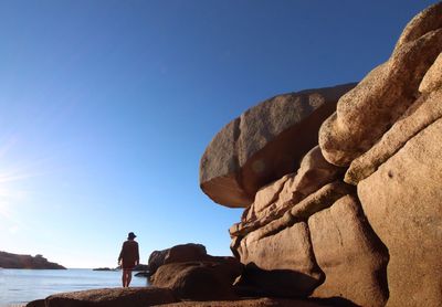 Rear view of woman standing by rock formation at sea against clear blue sky