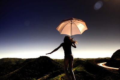 Rear view of young woman with umbrella standing on mountain against clear sky during sunset