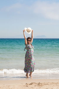 Portrait of woman with arms raised holding hat while standing at beach