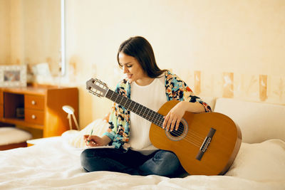 Young woman holding guitar writing on notebook while sitting on bed