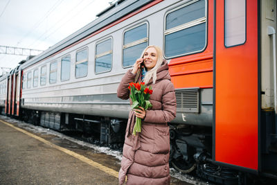 Joyful girl at the train station with flowers talking on the phone