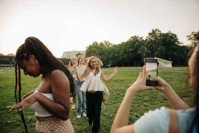 Teenage girl photographing female friends enjoying dance in park at sunset