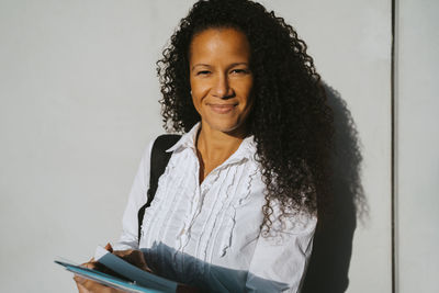 Portrait of smiling woman with curly hair holding book against wall at college campus