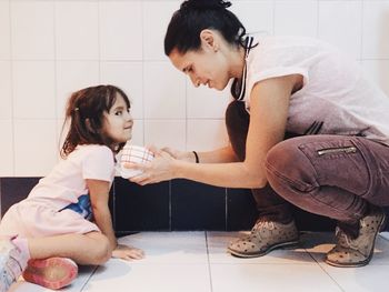 Side view full length of mother with daughter holding bowl by wall on floor