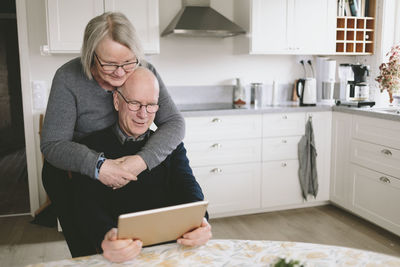 Smiling couple embracing and using tablet at kitchen table