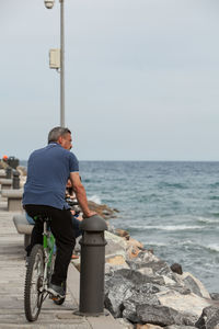 Rear view of man riding bicycle on footpath by sea against sky