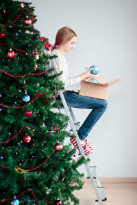 Teenage girl looking at christmas ornament while sitting on ladder by christmas tree