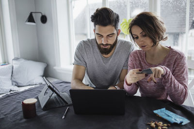 Woman holding credit card while man using laptop on table at home