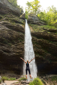 Rear view of woman with arms raised standing against waterfall