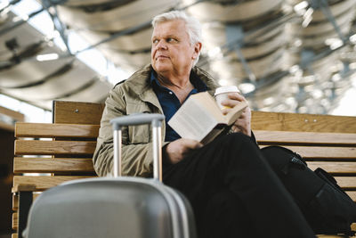 Low angle view of senior businessman with book and disposable cup sitting by suitcase on bench at food court in railroad