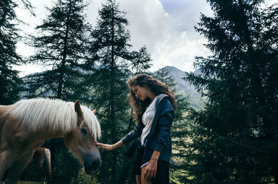 Young woman touching horse while standing against trees