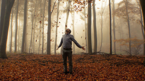 Dry leaves falling on man standing in forest with arms outstretched during foggy weather