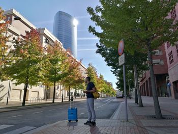 Man with suitcase standing by trees in city against sky