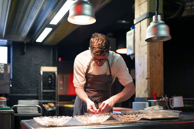 Young serious chef man in cooking apron serving meal in dishes on restaurant kitchen