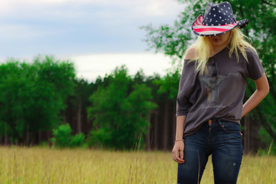 Country girl with forest behind her wearing an american flag hat standing in a prairie