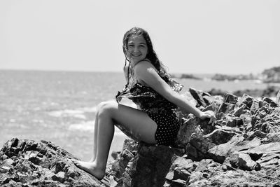 Side view portrait of smiling woman sitting on rocks at beach against sky