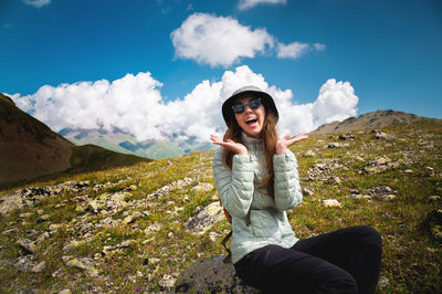 Young woman traveler sits on a rock against the backdrop of mountains and fluffy clouds with a blue
