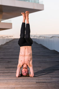 Full length of shirtless man doing headstand on boardwalk