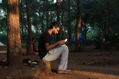 Side view of young man reading book while sitting in forest