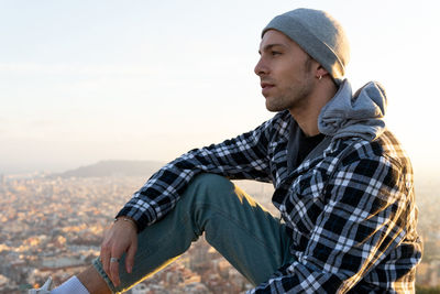 Side view of thoughtful hipster male traveler in warm checkered hoodie and hat looking away while sitting on hill against blurred town in autumn day