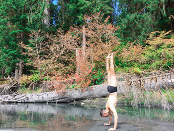 Side view of shirtless man practicing handstand on riverbank against trees in forest during autumn