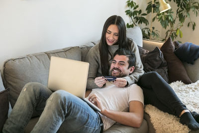 Happy couple using credit card while shopping online through laptop on sofa at home