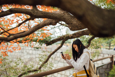 Portrait of young woman taking photo using mobile phone under a tree at the riverside 