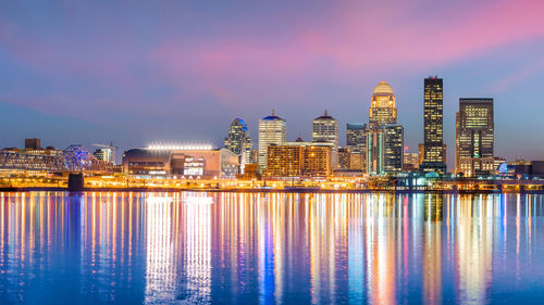 Illuminated buildings by river against sky at night