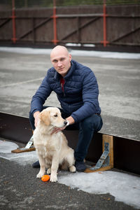 Portrait of man with golden retriever sitting on small fence during winter