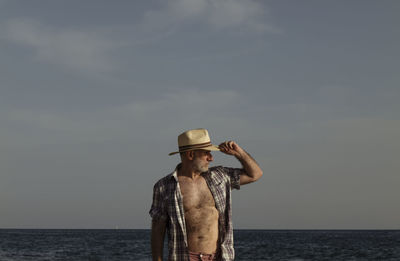 Adult man in hat with open shirt on beach against sea and sky