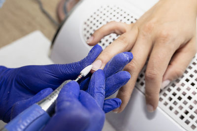 The manicurist grinds the nail plate with a milling machine in blue latex gloves.