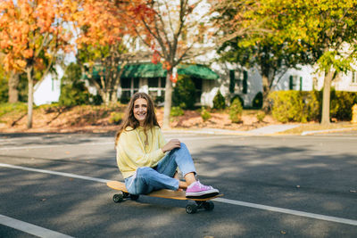 Smiling girl sitting on longboard in autumn weather in a neighborhood