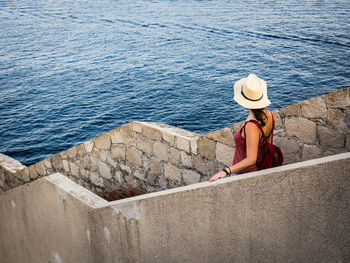 Rear view of woman sitting on retaining wall by sea