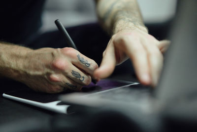 Crop guy in casual outfit with tattoos sketching on computer in bright tattoo studio near tattooist making tattoo for client