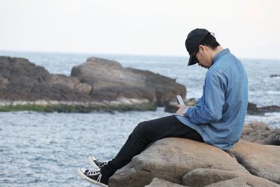 Side view of young man using phone while sitting on rock at beach