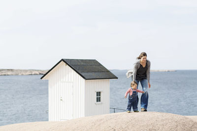 Mother and baby girl walking at beach by beach cabin against clear sky