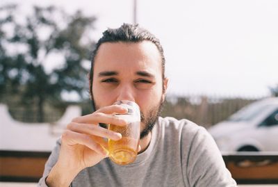 Portrait of young man drinking beer while sitting against sky