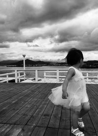 Small girl walking on pier against cloudy sky