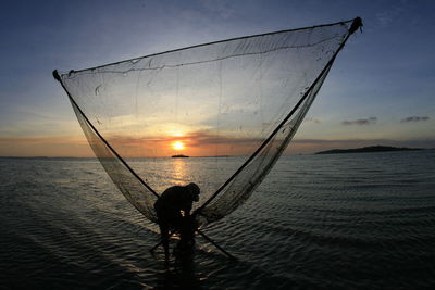 Fisherman working on shore at beach against sky during sunset