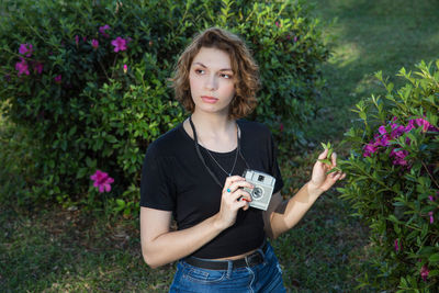 Beautiful young woman holding camera while standing by plants