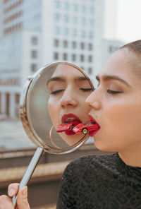 Close-up of young woman with red toy car in mouth while holding mirror with reflection in city