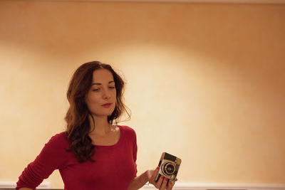 Woman holding camera while standing against wall