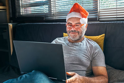 Handsome smiling mature man wearing reading glasses and christmas red hat has video chat on laptop.