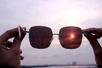 Midsection of person holding sunglasses against sky during sunset