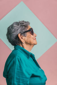 Side view of happy modern aged female with gray hair and in trendy sunglasses on pink and blue background in studio and looking away