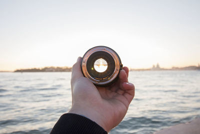 Cropped hand holding lens against sea during sunset