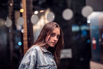 Portrait of woman standing against illuminated city seen through glass window at night