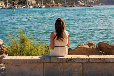 Rear view of woman sitting on stone wall by sea