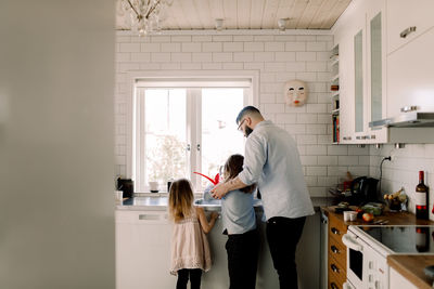 Man standing with girl while assisting daughter at kitchen sink