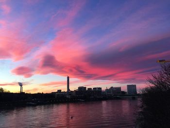 Silhouette buildings by river against sky at sunset